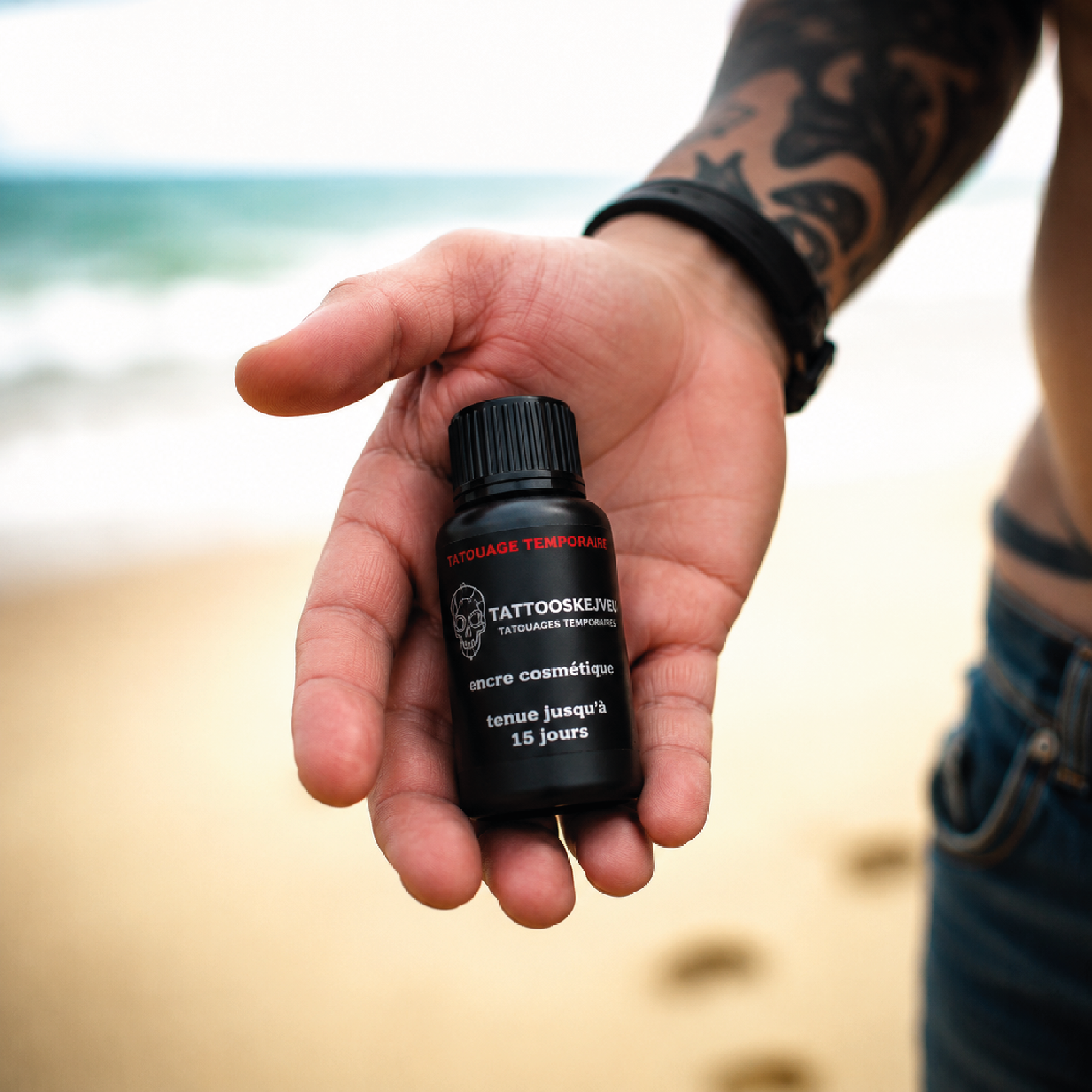 Main tenant un flacon d’encre noire pour tatouage temporaire avec tenue jusqu’à 15 jours sur fond de plage.